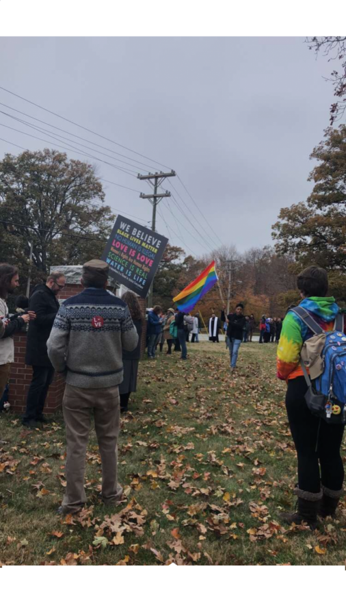 A rainbow flag representing the LGBTQIA community is hoisted by an individual walking along the Barrier of Love, while another individual holds up a sign that says, We believe Black Lives Matter, No Human Is Illegal, Love is Love, Womens Rights Are Human Rights, Science is Real, Water is Life.